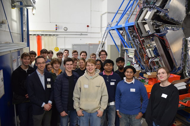 A group of persons stands in a large room. At the right-hand corner a huge experiment setup is visible. A group of persons stands in a large room. At the right-hand corner a huge experiment setup is visible.