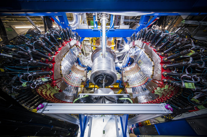 Zwei silbern glitzernde Halbkugeln stehen zu den Seiten eines silbernen Zylinders. Viele Kabel führen an den Seiten weg.