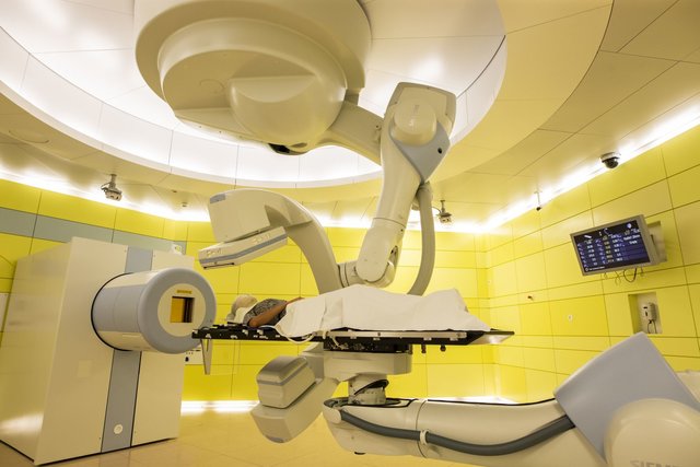 A person is positioned on a patient bench, held by a robot, in front of a cylindrical beam exit window. A person is positioned on a patient bench, held by a robot, in front of a cylindrical beam exit window.