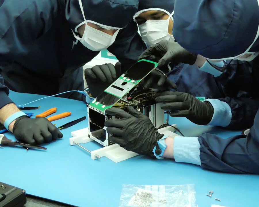 Mehrere Personen in Schutzanzügen beugen sich über einen rechteckigen Kasten mit Elektronik.